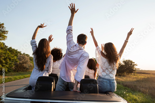 Back view. Young guys are sitting and holding hands up in a black cabriolet on the country road on a sunny day.