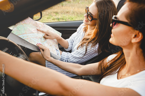 Two dark-haired young women in sunglasses are sitting in a black cabriolet on a sunny day. One of them keeps map in her hands. © Leika production