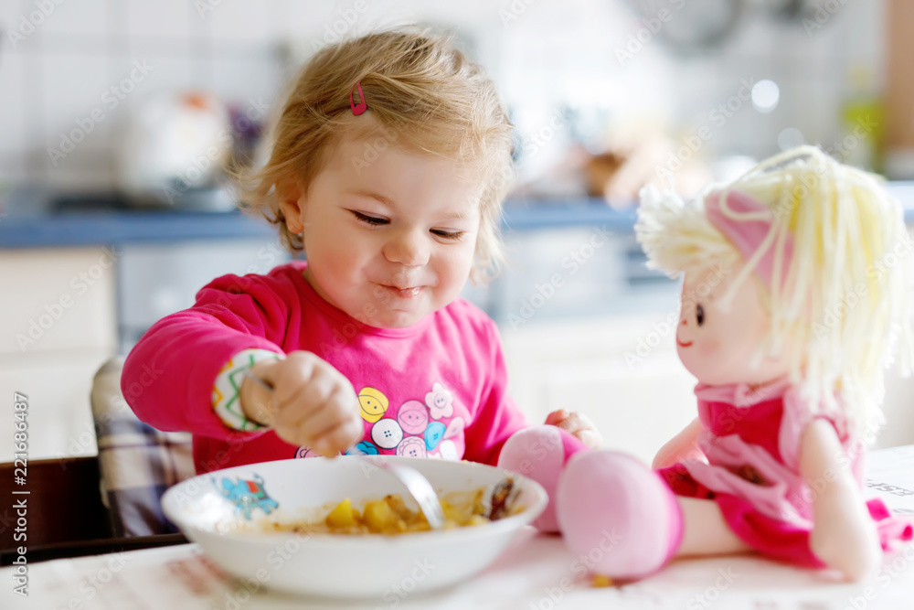 Adorable baby girl eating from fork vegetables and pasta. Little child feeding and playing with toy doll. Cute toddler, daughter with spoon sitting in highchair and learning to eat by itself. Adorable baby girl eating from fork vegetables and pasta. Little child feeding and playing with toy doll. Cute toddler, daughter with spoon sitting in highchair and learning to eat by itself.