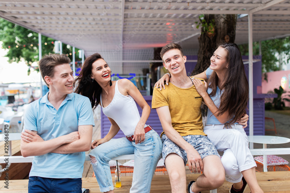 A company of good-looking friends is laughing and sitting on the railing in the nice summer cafe. Entertainment, having good time. Friendship. A company of good-looking friends is laughing and sitting on the railing in the nice summer cafe. Entertainment, having good time. Friendship.