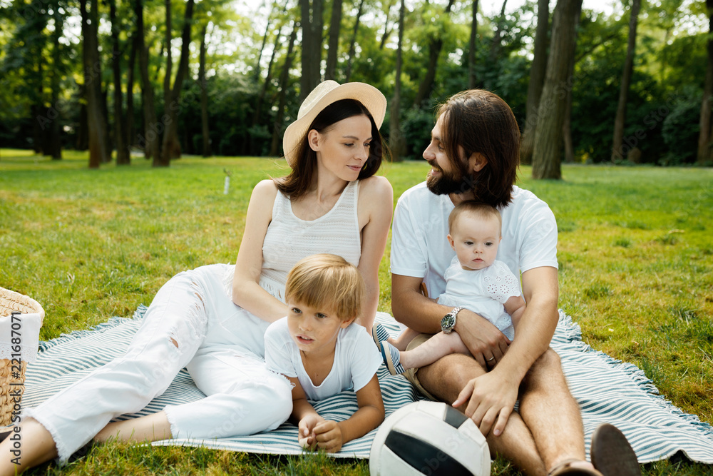 Beautiful young family dressed in white clothes is sitting on a blanket on the lawn on a warm sunny day in the park. Beautiful young family dressed in white clothes is sitting on a blanket on the lawn on a warm sunny day in the park.