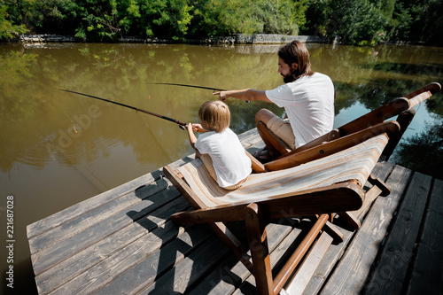Young dark-haired father and his little son are sitting in recliners on the wooden pier with fishing rods and fishing.
