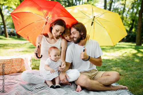 Cheerful parents with two kids have a rest on the lawn under the bright red and yellow umbrellas covering them from the sun.