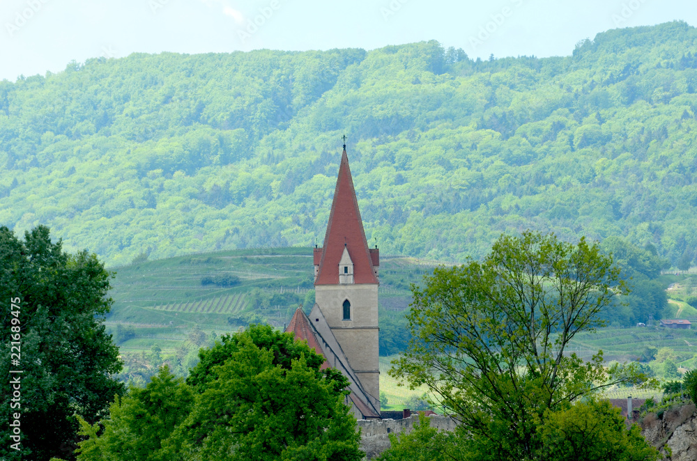 The red steeple of a church is seen against forest-covered hills. A wall with a guard tower is in front of the church. The sky is blue.