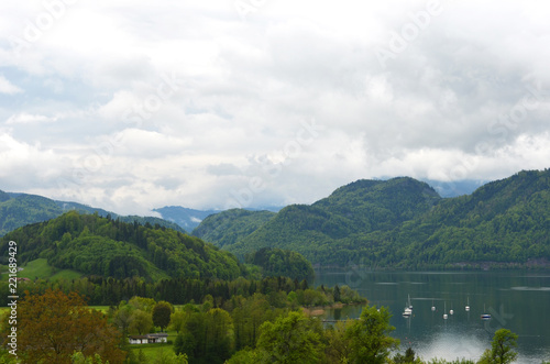 Forest covered hills surround a lake. Some yachts are on the water, with a house in the foreground. The sky is overcast..