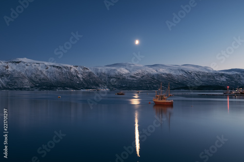 Winter landscape of a Norwegian village in winter with a fishing boat at the forground. Moon over the fjord in Norway.