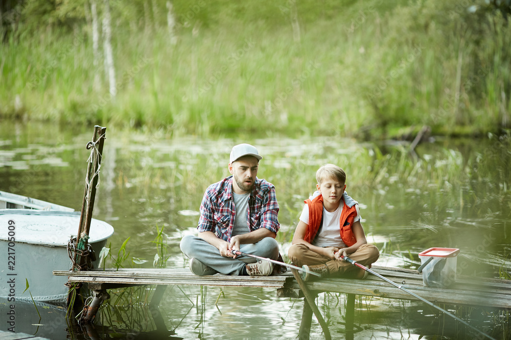 Father and son sitting on pontoon with their rods in water and waiting for fish Father and son sitting on pontoon with their rods in water and waiting for fish