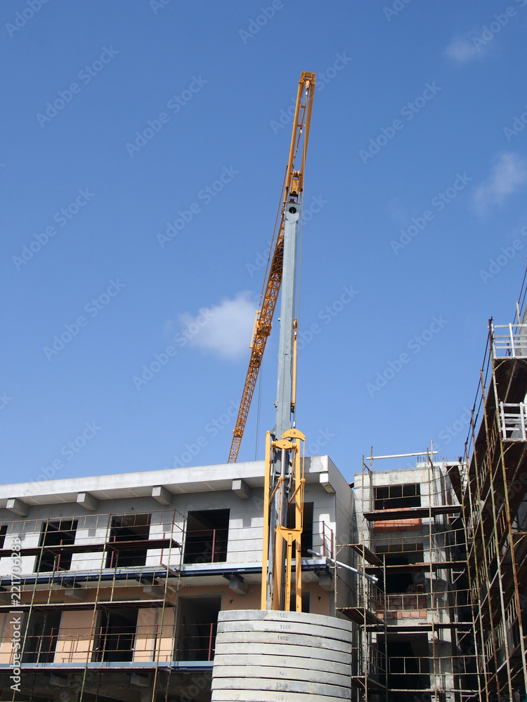 A yellow construction crane working on a modern concrete apartment development with scaffolding with blue sky and sunlight