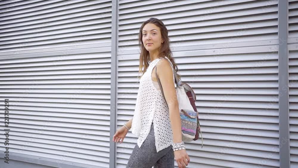slim cheerful student woman is walking outdoors in city along buildings in daytime, turning to camera slim cheerful student woman is walking outdoors in city along buildings in daytime, turning to camera