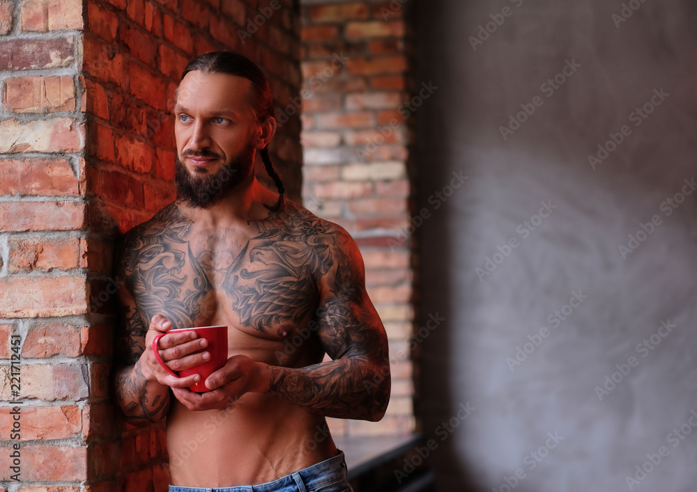 Stylish shirtless bearded male with muscular and tattooed body holds cup of coffee while leaning on a wall and looks out the window.
