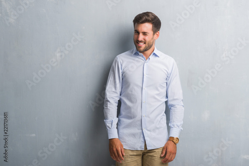 Handsome young business man over grey grunge wall wearing elegant shirt winking looking at the camera with sexy expression, cheerful and happy face.