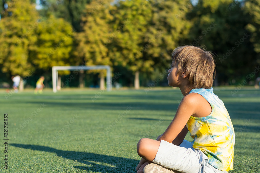 A boy is sitting on the grass at the edge of a stadium and watching football.