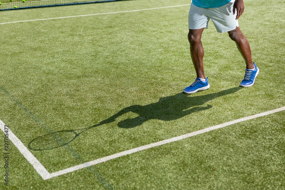 Sporty man is spending time on court outdoor on sunny day. He is moving while holding racket while casting shadow on playground. Copy space in left side
