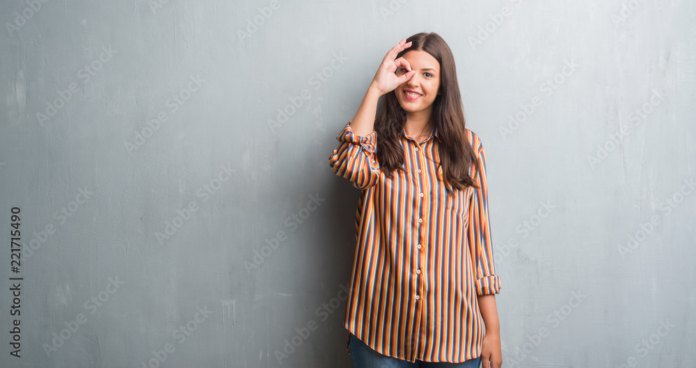 Young brunette woman over grunge grey wall doing ok gesture with hand smiling, eye looking through fingers with happy face. Young brunette woman over grunge grey wall doing ok gesture with hand smiling, eye looking through fingers with happy face.