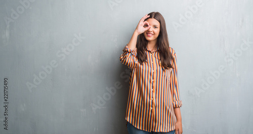Young brunette woman over grunge grey wall doing ok gesture with hand smiling  eye looking through fingers with happy face.