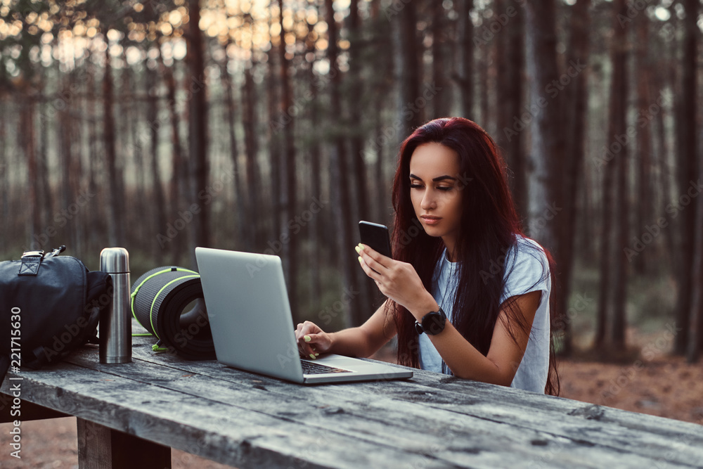 Portrait of a hipster girl using a smartphone while sitting on a wooden bench with open laptop in the beautiful autumn forest. Portrait of a hipster girl using a smartphone while sitting on a wooden bench with open laptop in the beautiful autumn forest.