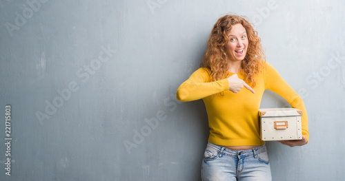 Young redhead woman over grey grunge wall holding box very happy pointing with hand and finger
