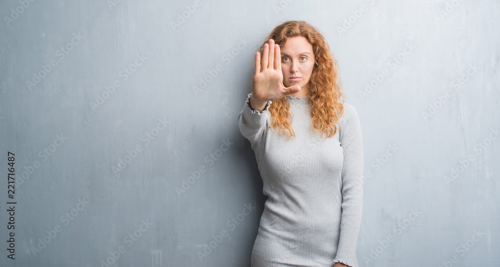 Young redhead woman over grey grunge wall with open hand doing stop sign with serious and confident expression, defense gesture Young redhead woman over grey grunge wall with open hand doing stop sign with serious and confident expression, defense gesture