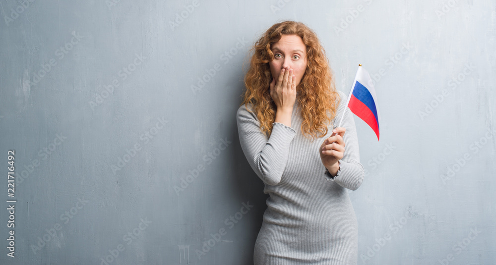 Young redhead woman over grey grunge wall holding flag of Russia cover mouth with hand shocked with shame for mistake, expression of fear, scared in silence, secret concept Young redhead woman over grey grunge wall holding flag of Russia cover mouth with hand shocked with shame for mistake, expression of fear, scared in silence, secret concept