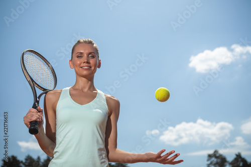 Low angle of grinning woman preparing for playing match under blue sky. She is standing and holding racket while putting it on shoulder. Girl is throwing up ball with hand © Yakobchuk Olena