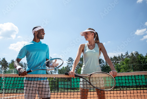 Smiling friends are having double set on court on sunny warm day. Man and woman are standing near net with rackets and discussing following match. Enjoying active games with close people concept © Yakobchuk Olena