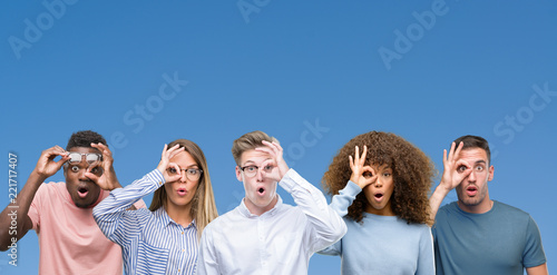 Composition of group of friends over blue blackground doing ok gesture shocked with surprised face, eye looking through fingers. Unbelieving expression.