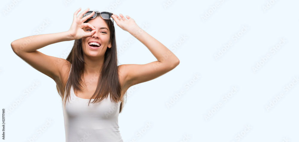 Young beautiful hispanic wearing sunglasses doing ok gesture with hand smiling, eye looking through fingers with happy face. Young beautiful hispanic wearing sunglasses doing ok gesture with hand smiling, eye looking through fingers with happy face.