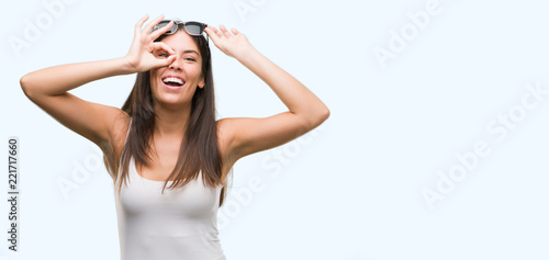 Young beautiful hispanic wearing sunglasses doing ok gesture with hand smiling, eye looking through fingers with happy face.