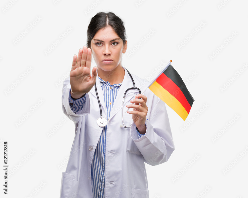 Young hispanic doctor woman holding flag of germany with open hand doing stop sign with serious and confident expression, defense gesture Young hispanic doctor woman holding flag of germany with open hand doing stop sign with serious and confident expression, defense gesture