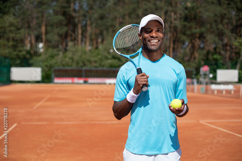 Waist up portrait of cheerful guy standing on sunny court. He is holding racket and ball in his hands while preparing for game. Copy space in left side © Yakobchuk Olena