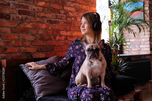 Happy blonde woman in dress sitting with her cute pug on a handmade sofa and listening to music in a room with loft interior. © Fxquadro