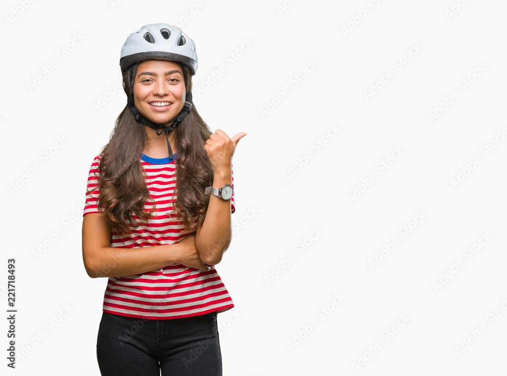 Young arab cyclist woman wearing safety helmet over isolated background smiling with happy face looking and pointing to the side with thumb up. Young arab cyclist woman wearing safety helmet over isolated background smiling with happy face looking and pointing to the side with thumb up.