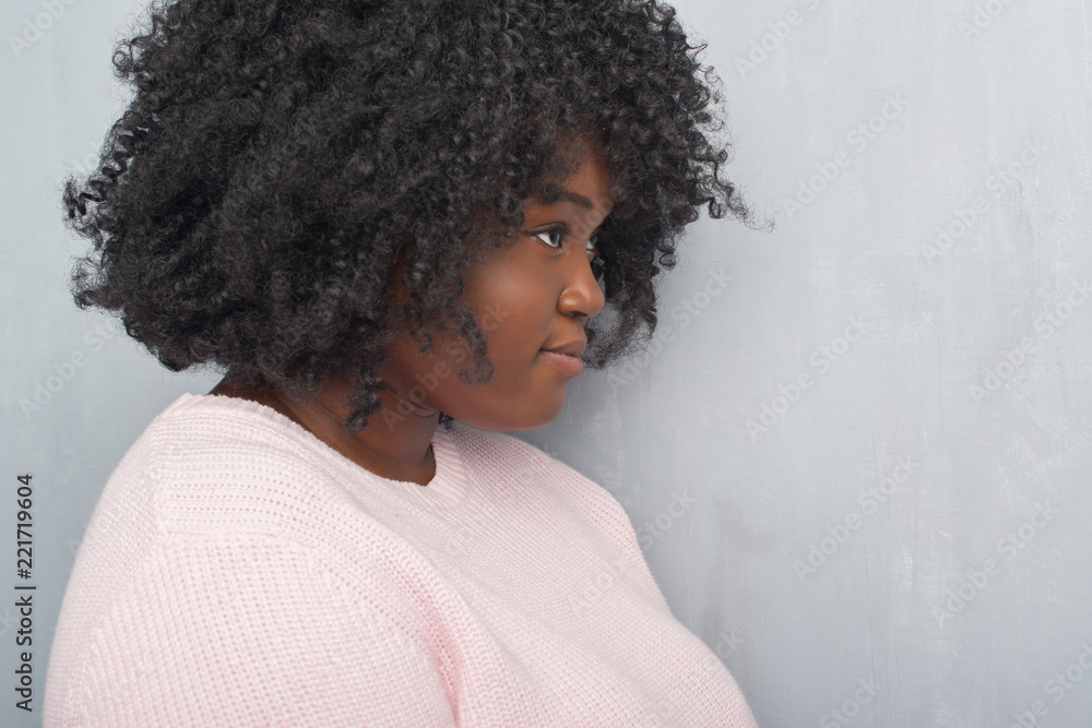 Young african american plus size woman over grey grunge wall wearing winter sweater looking to side, relax profile pose with natural face with confident smile. Young african american plus size woman over grey grunge wall wearing winter sweater looking to side, relax profile pose with natural face with confident smile.