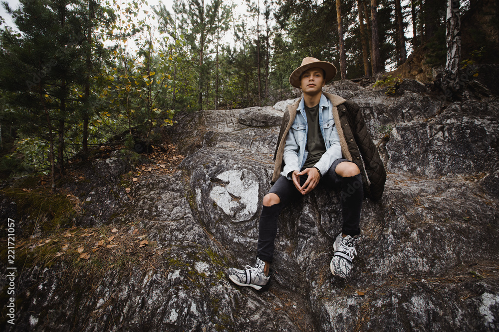 Naklejka premium Young handsome man in jeans and warm jacket sitting on rocky cliff in forest on background of green trees