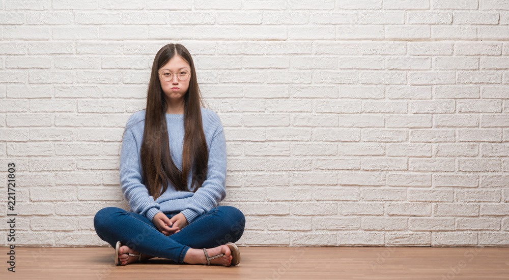 Young Chinese woman sitting on the floor over brick wall puffing cheeks with funny face. Mouth inflated with air, crazy expression. Young Chinese woman sitting on the floor over brick wall puffing cheeks with funny face. Mouth inflated with air, crazy expression.