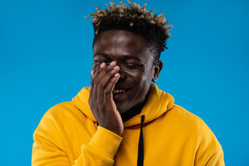 Stop me. Close up portrait of cheerful guy with closed eyes covering face with hand. He is wearing yellow hoodie and standing on blue background