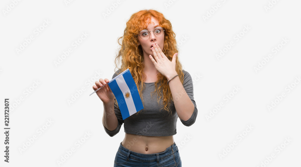 Young redhead woman holding flag of Argentina cover mouth with hand shocked with shame for mistake, expression of fear, scared in silence, secret concept Young redhead woman holding flag of Argentina cover mouth with hand shocked with shame for mistake, expression of fear, scared in silence, secret concept