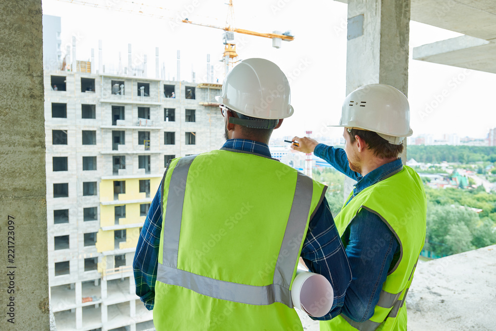 Rear view of constriction workers in hardhats and green vests discussing building structure, male engineer pointing at building under construction Rear view of constriction workers in hardhats and green vests discussing building structure, male engineer pointing at building under construction
