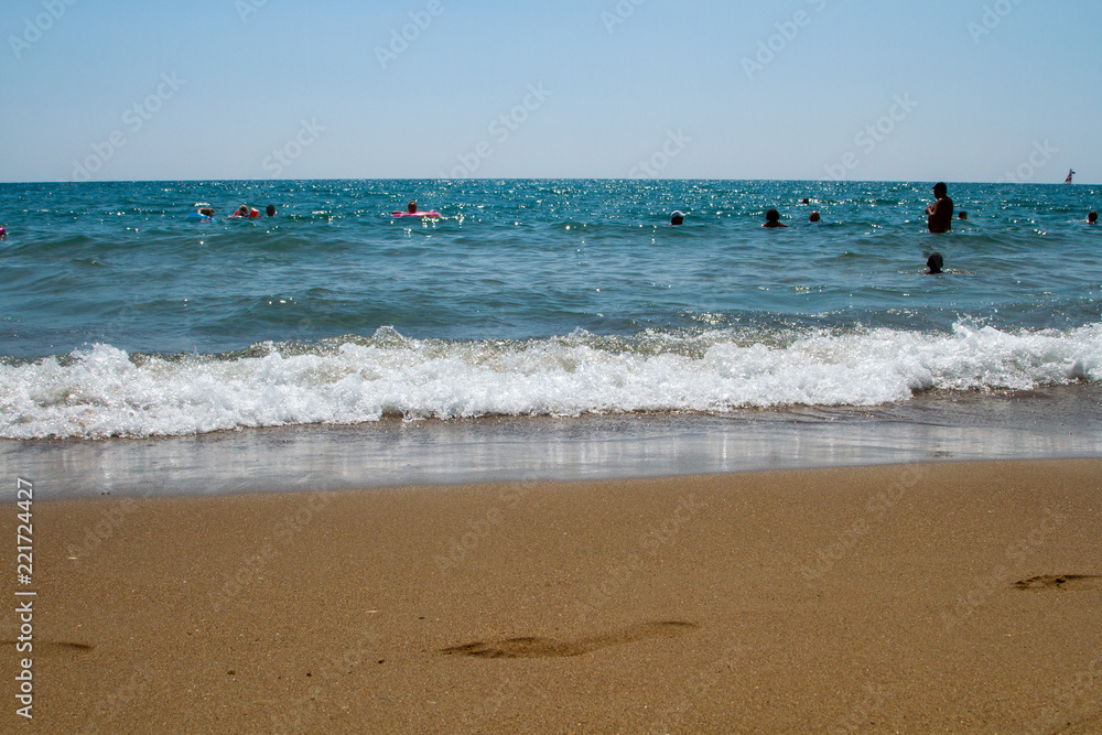 Sea beach with bathing people, sea wave, surf, and with human footprints in the sand on a bright sunny day.