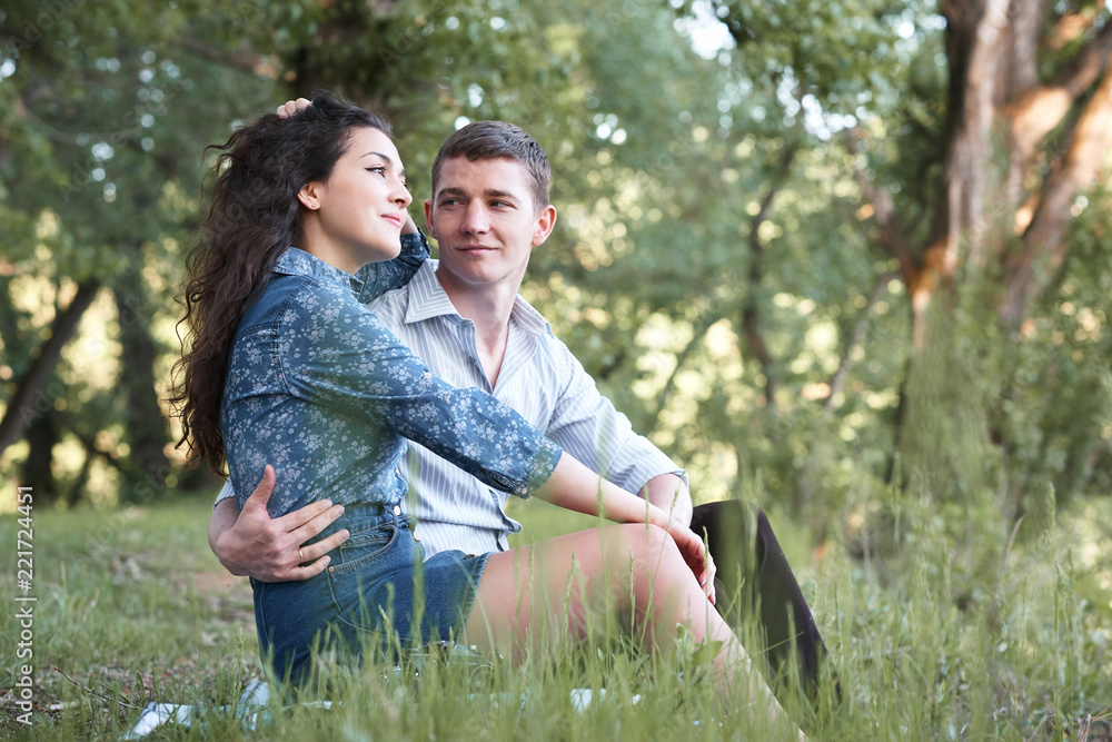 young couple sitting on the grass in the forest and looking on sunset, summer nature, bright sunlight, shadows and green leaves, romantic feelings young couple sitting on the grass in the forest and looking on sunset, summer nature, bright sunlight, shadows and green leaves, romantic feelings