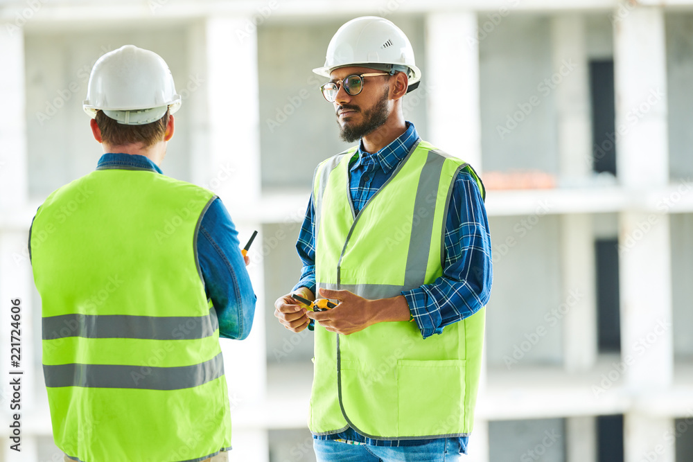 Serious confident middle-eastern worker in glasses and hardhat attentively listening to colleagues while they standing at apartment block site Serious confident middle-eastern worker in glasses and hardhat attentively listening to colleagues while they standing at apartment block site