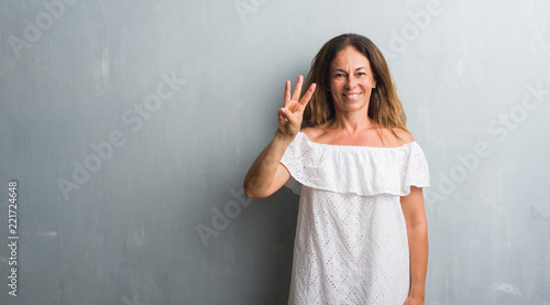 Middle age hispanic woman standing over grey grunge wall showing and pointing up with fingers number three while smiling confident and happy.