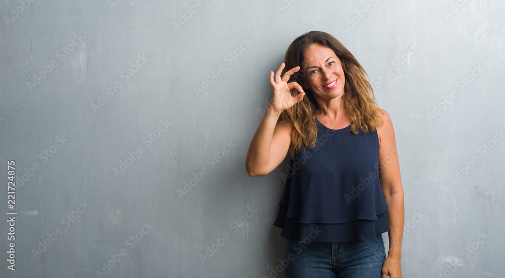 Middle age hispanic woman standing over grey grunge wall smiling positive doing ok sign with hand and fingers. Successful expression. Middle age hispanic woman standing over grey grunge wall smiling positive doing ok sign with hand and fingers. Successful expression.