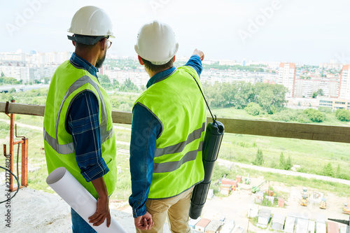 Rear view of construction engineers in hardhats and vests contemplating next residential district and discussing new building project, man with tube for blueprints pointing with finger at buildings photo