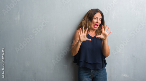 Middle age hispanic woman standing over grey grunge wall afraid and terrified with fear expression stop gesture with hands, shouting in shock. Panic concept.