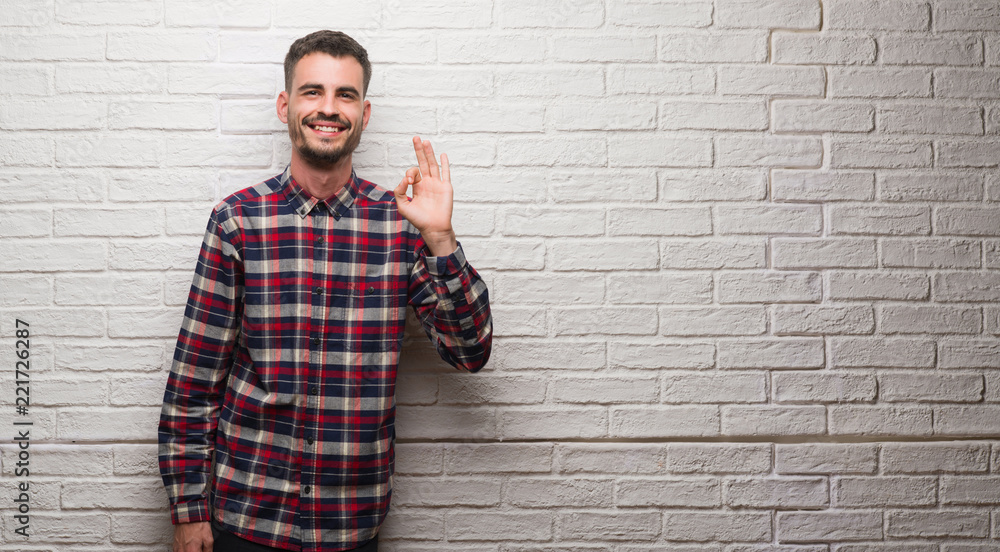 Young adult man over white brick wall doing ok sign with fingers, excellent symbol Young adult man over white brick wall doing ok sign with fingers, excellent symbol