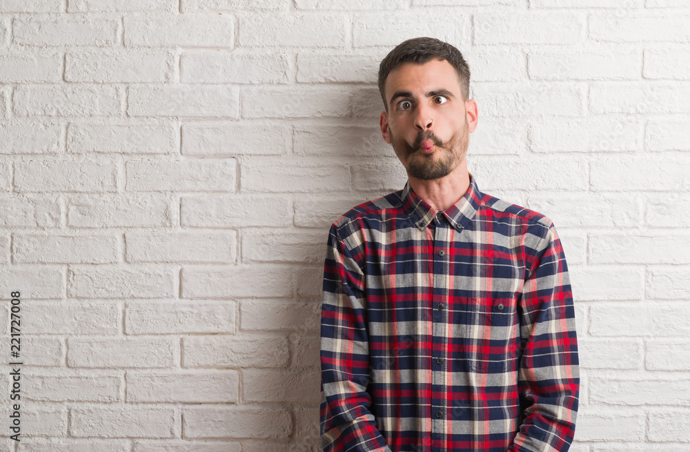 Young adult man standing over white brick wall making fish face with lips, crazy and comical gesture. Funny expression. Young adult man standing over white brick wall making fish face with lips, crazy and comical gesture. Funny expression.
