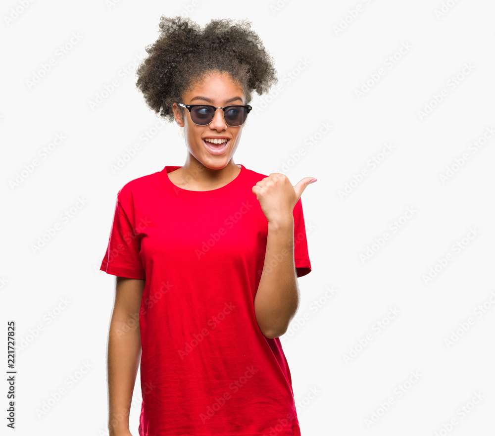 Young afro american woman wearing sunglasses over isolated background smiling with happy face looking and pointing to the side with thumb up. Young afro american woman wearing sunglasses over isolated background smiling with happy face looking and pointing to the side with thumb up.