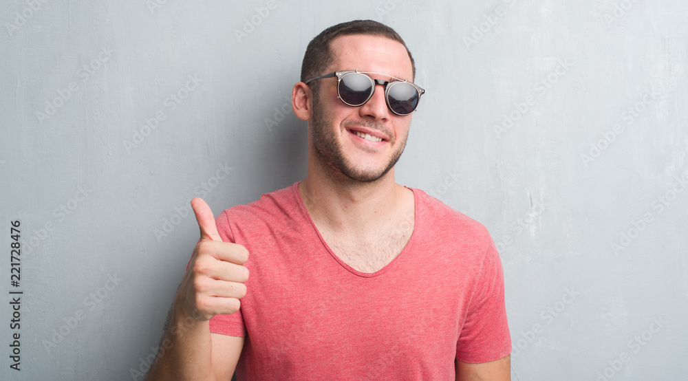Naklejka premium Young caucasian man over grey grunge wall wearing sunglasses happy with big smile doing ok sign, thumb up with fingers, excellent sign