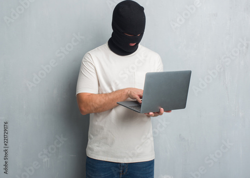 Young caucasian hacker man over grey grunge wall doing cyber attack using laptop with a confident expression on smart face thinking serious